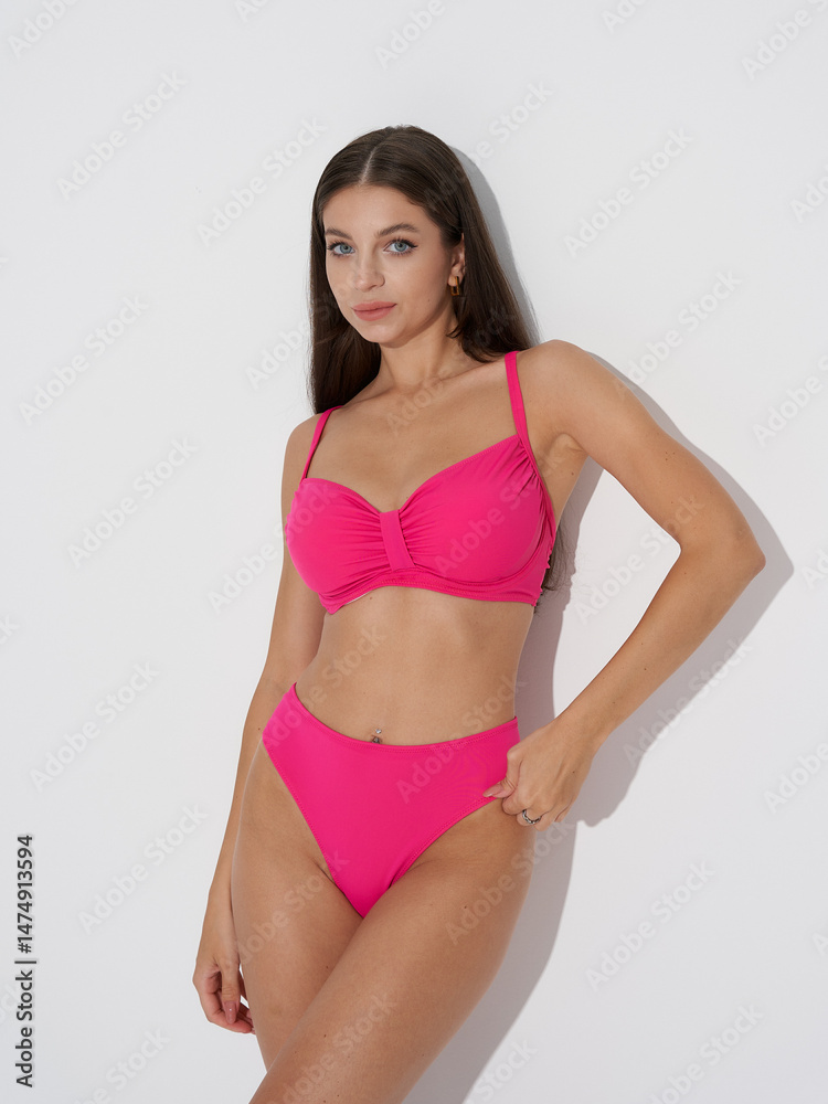 Fototapeta premium Brunette woman wearing a bright pink bikini poses confidently against a plain backdrop. She has long hair, earrings, and a ring on her finger, exuding elegance and style