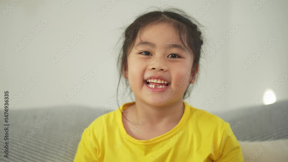 Happy young girl in yellow shirt sitting on sofa with calm expression, enjoying leisure time at home, looking directly at camera with curious eyes Happy young girl in yellow shirt sitting on sofa with calm expression, enjoying leisure time at home, looking directly at camera with curious eyes