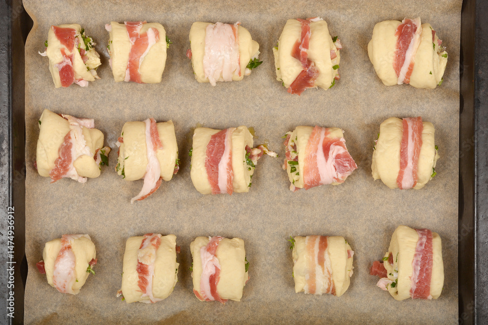 Bacon-wrapped dough rolls filled with a mixture of diced pancetta and herbs are arranged on a baking sheet, ready to be baked