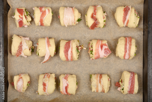 Bacon-wrapped dough rolls filled with a mixture of diced pancetta and herbs are arranged on a baking sheet, ready to be baked