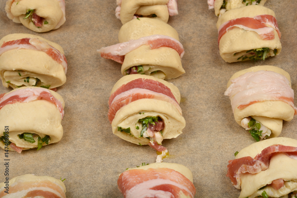 Bacon-wrapped dough rolls filled with a mixture of diced pancetta and herbs are arranged on a baking sheet, ready to be baked