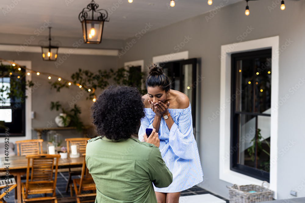 Naklejka premium Man proposing to surprised woman on patio, romantic moment under string lights