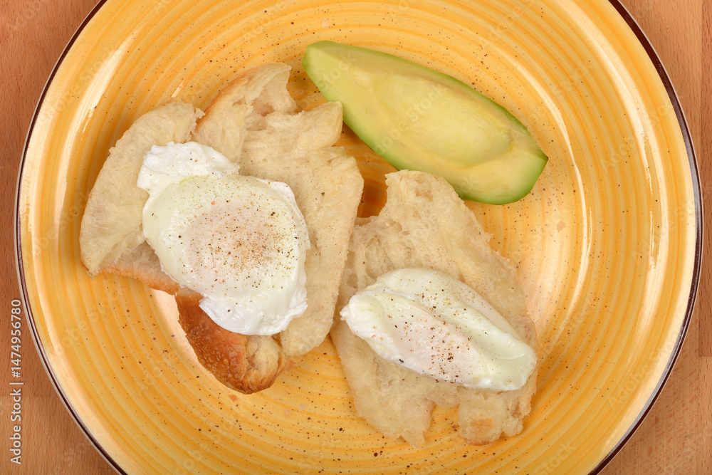 A top-down view shows two poached eggs seasoned with pepper on pieces of bread, accompanied by a slice of avocado on a yellow plate