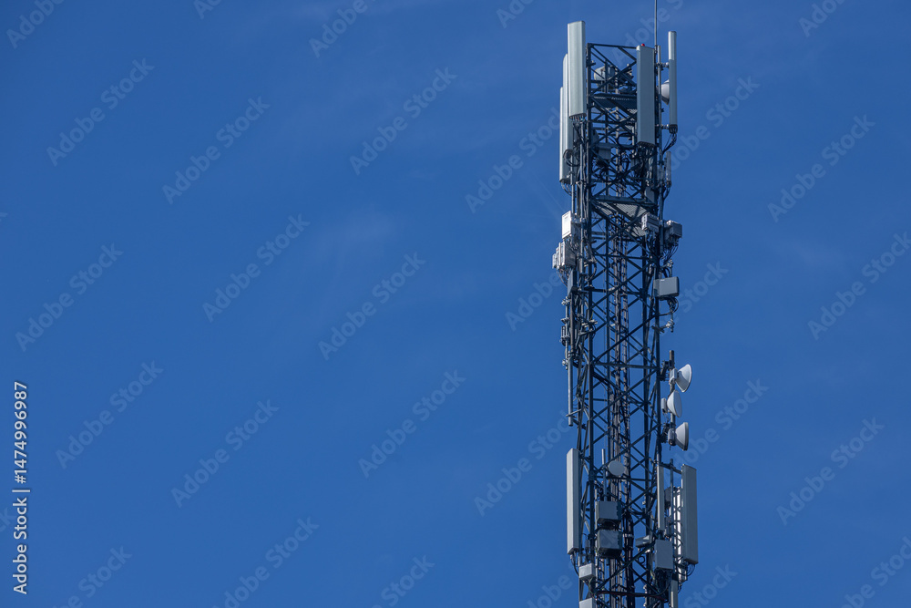 A tall communications tower equipped with antennas and satellite dishes stands sharply against a vivid blue sky, symbolizing modern technology and connectivity. Includes copy space. A tall communications tower equipped with antennas and satellite dishes stands sharply against a vivid blue sky, symbolizing modern technology and connectivity. Includes copy space.