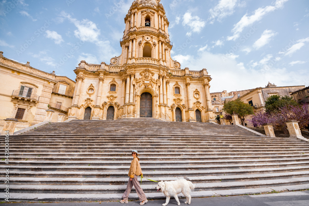 Fototapeta premium Woman walks her large white dog past the grand staircase of the Cathedral of San Giorgio in Modica, Sicily, under a bright blue sky