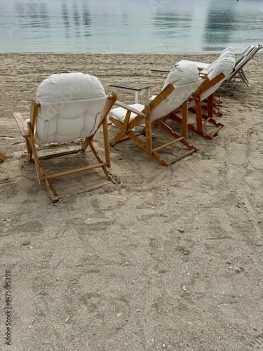 Wooden beach chairs with white cushions, lined up on sandy shore, overlooking calm, reflective waters under a serene sky.