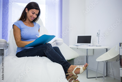 Woman is writing on blue clipboard seated on white treatment chair in clinic room with laptop