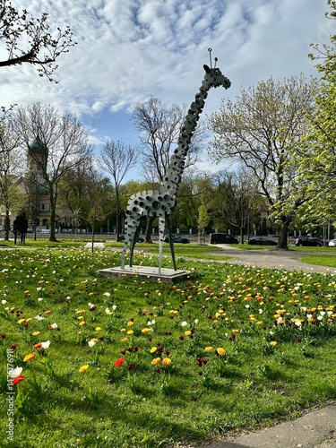 Decorative metal giraffe sculpture standing in a vibrant tulip flowerbed in a park on a sunny day. The background features a clear blue sky, creating a cheerful and artistic spring scene.