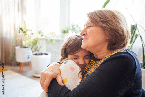 A smiling elderly woman hugs her sweet granddaughter tightly. A little girl in casual clothes snuggles up to her grandmother.