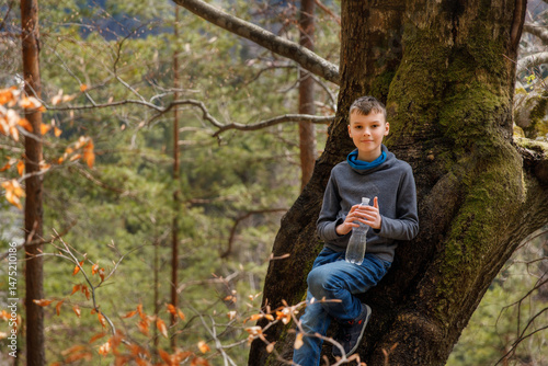  A young, fair-haired boy in a grey sweater and jeans sits comfortably against a large tree covered in green moss, holding a clear water bottle.