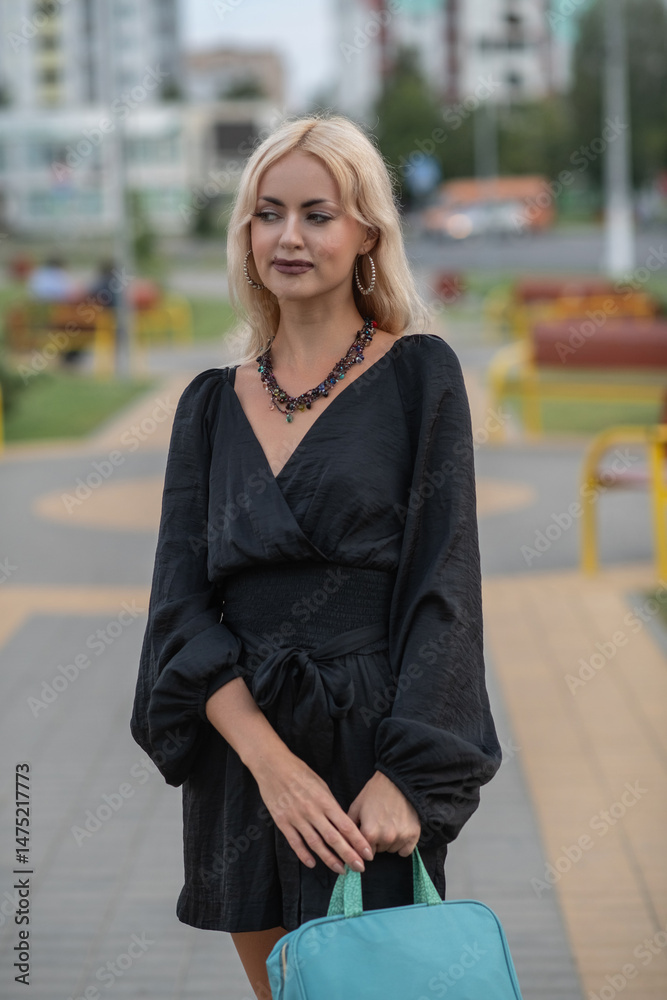 Fototapeta premium Portrait of a young beautiful blonde girl in a dark dress in a city dress in the summer.