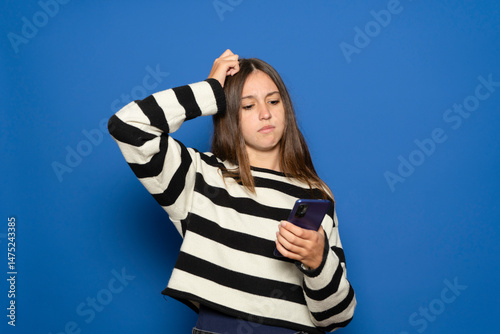 Hispanic teenage girl about 15 years old wearing striped sweater scratching her head with questioning expression and holding a smartphone over blue background.