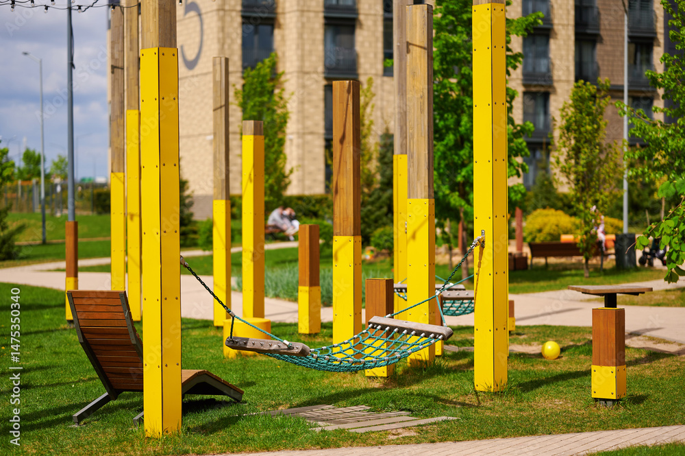 Fototapeta premium modern playground with yellow structures and net hammock provides urban oasis amid apartment buildings. green lawn and benches create vibrant community gathering space