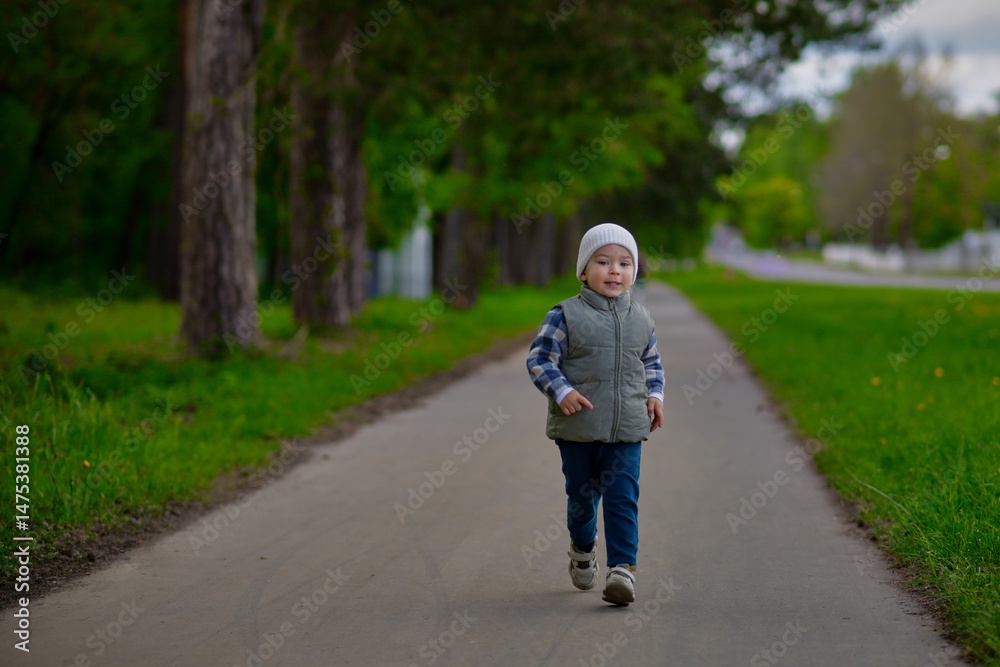 Fototapeta premium Happy little boy running on a forest path in spring, wearing a warm vest and beanie. Active child enjoying nature and freedom in a green park on a fresh outdoor day.