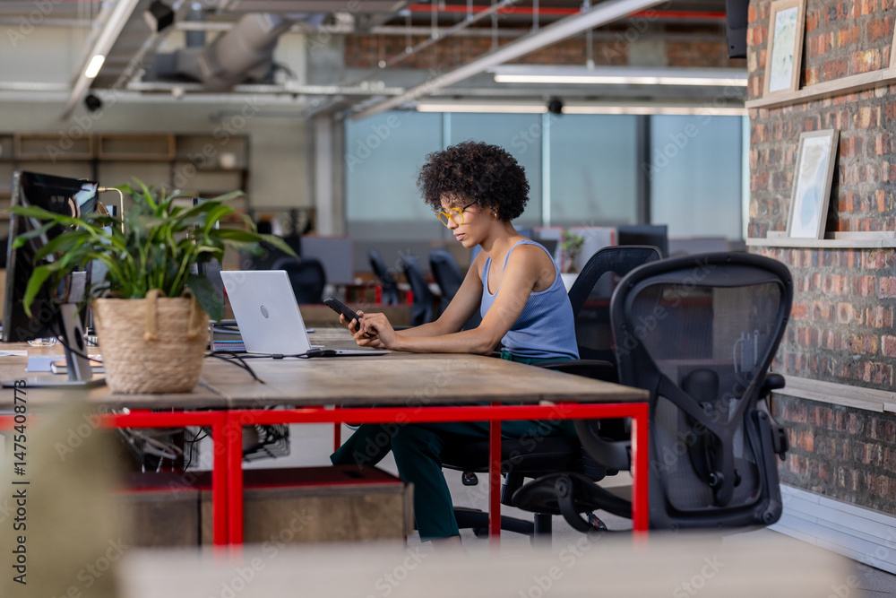 Fototapeta premium African American woman using smartphone and laptop at modern office desk