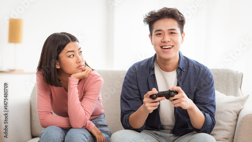 Displeased Japanese Girlfriend Looking At Infantile Boyfriend While He Playing Video Game Sitting On Sofa At Home. Male Emotional Immaturity, Gaming Addiction. Computer Games And Waste Of Time Concept