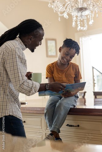 African American father and son reading book together in kitchen, sharing smiles