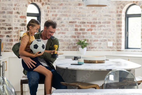 Father and daughter celebrating soccer victory at home, holding trophy and ball