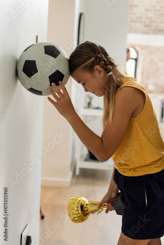 Girl holding trophy, leaning head on soccer ball at home, feeling thoughtful