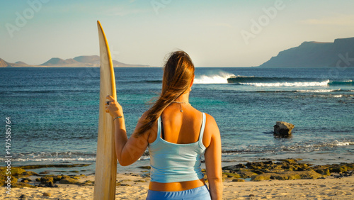 CLOSE UP: Surfer with wooden surfboard watches beautiful waves breaking in offshore wind. Young woman is checking out empty surf spot on Lanzarote island before heading for a morning surf session.