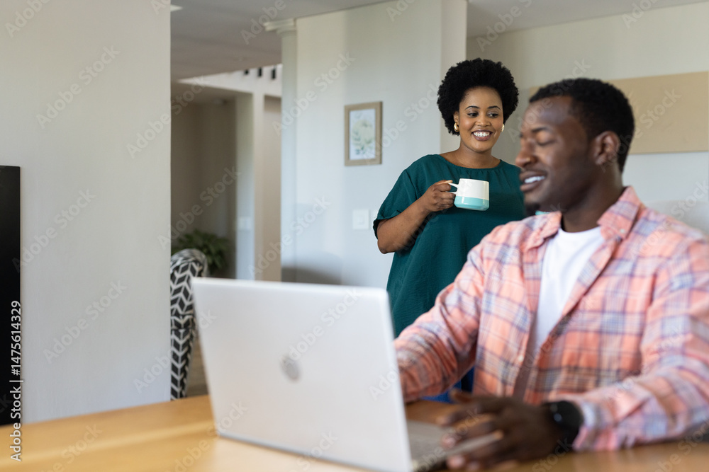Obraz premium Smiling woman holding coffee while man works on laptop at home
