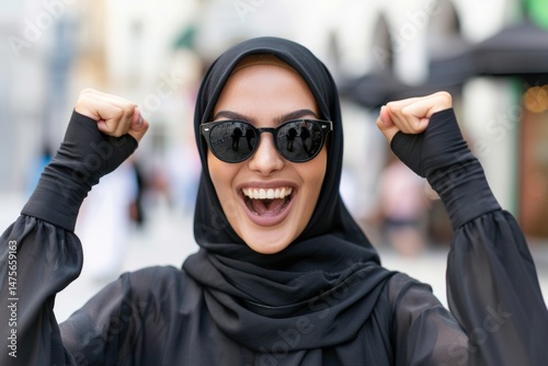Fashionable woman in black hijab and sunglasses expressing joy with raised fists in urban setting, showcasing happiness and celebration in city lifestyle.