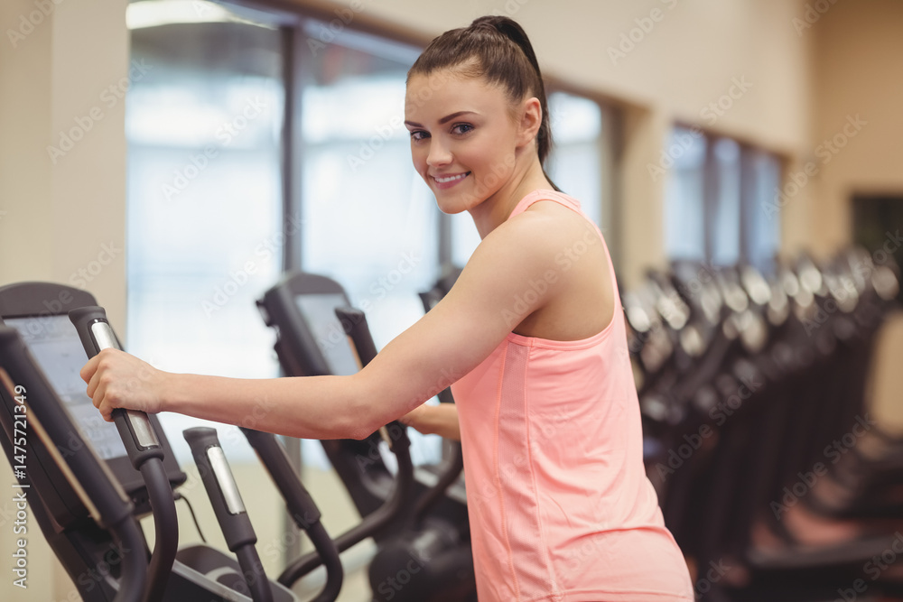 Fototapeta premium Exercising woman standing on elliptical in fitness center, with machine handles and mirror