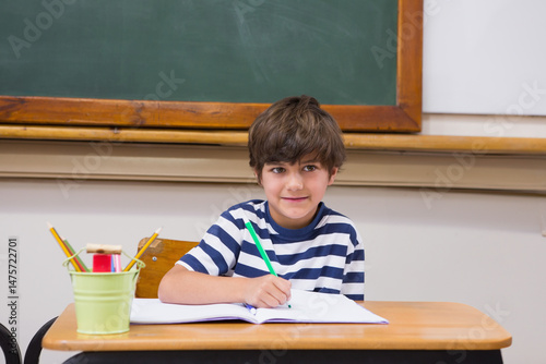 boy is writing in notebook with green pencil at school desk near green bucket in classroom