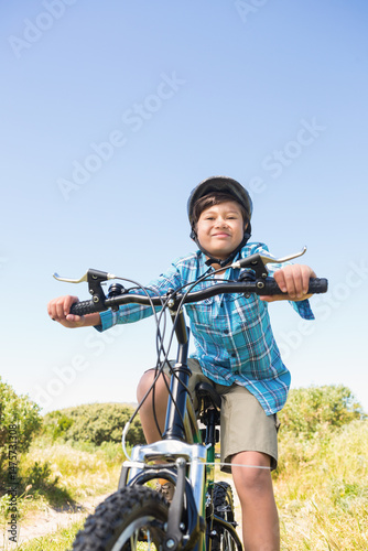 Boy wearing helmet and blue shirt riding mountain bike on grassy dirt trail among green shrubs