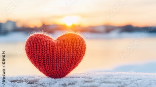 A soft red knitted heart stands on a snowy surface with a glowing sunset in the background, creating a peaceful winter atmosphere