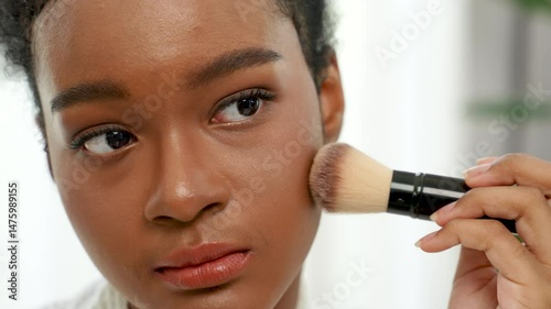 Beautiful black teenage girl using blusher to apply makeup in front of a mirror at home.