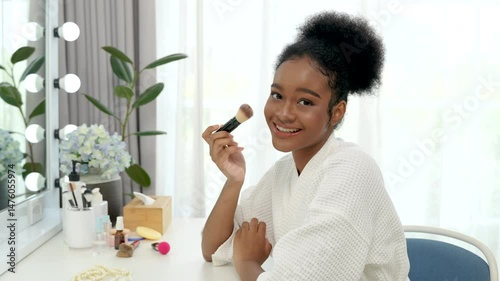Beautiful black teenage girl using blusher to apply makeup in front of a mirror at home.