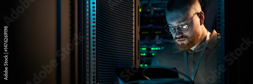 Young technician focusing on server maintenance in well-lit data center. Surrounded by networking equipment, performing checks on servers, concentrating intensely on task