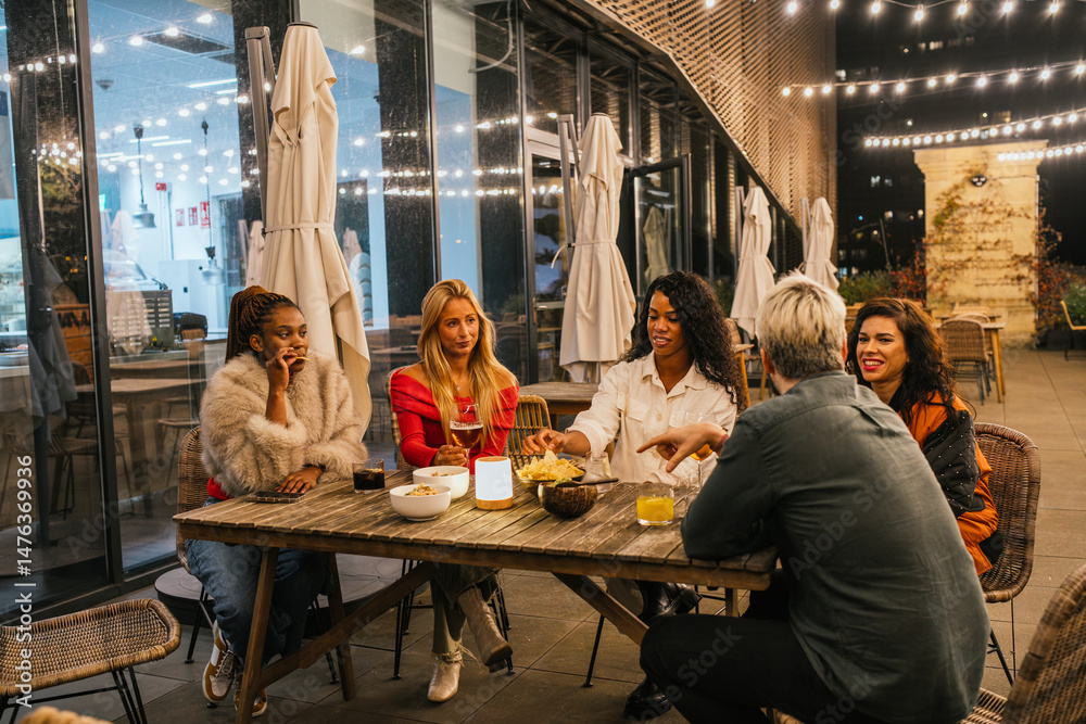 Naklejka premium Multiethnic group of friends having fun, eating, and drinking at a rooftop restaurant at night