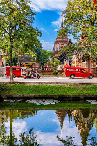 A scenic view of Chiang Mai city moat reflects the ancient Wat Lok Moli temple under a bright blue sky, with red songthaews and a tuk-tuk adding a touch of local life.