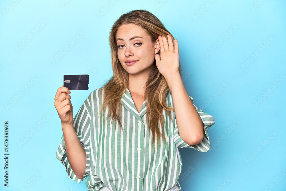 Young blonde with a credit card in blue studio trying to listening a gossip. Young blonde with a credit card in blue studio trying to listening a gossip.