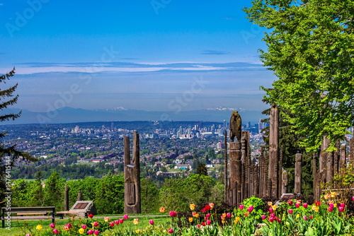 Totem poles in Burnaby Mountain Park on green grass next to trees and flower bed against blue sky, Vancouver skyline and mountain range on the horizon