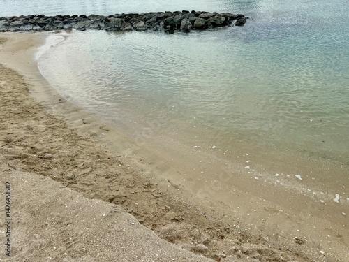 Calm beach shoreline with gentle waves, light sand, and a stone breakwater extending into the water. Horizontal