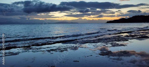 A breathtaking beach at sunset, with rugged rocks jutting from the shore. The sky glows with fiery oranges and pinks, reflecting off the calm waves in a tranquil, golden haze.
