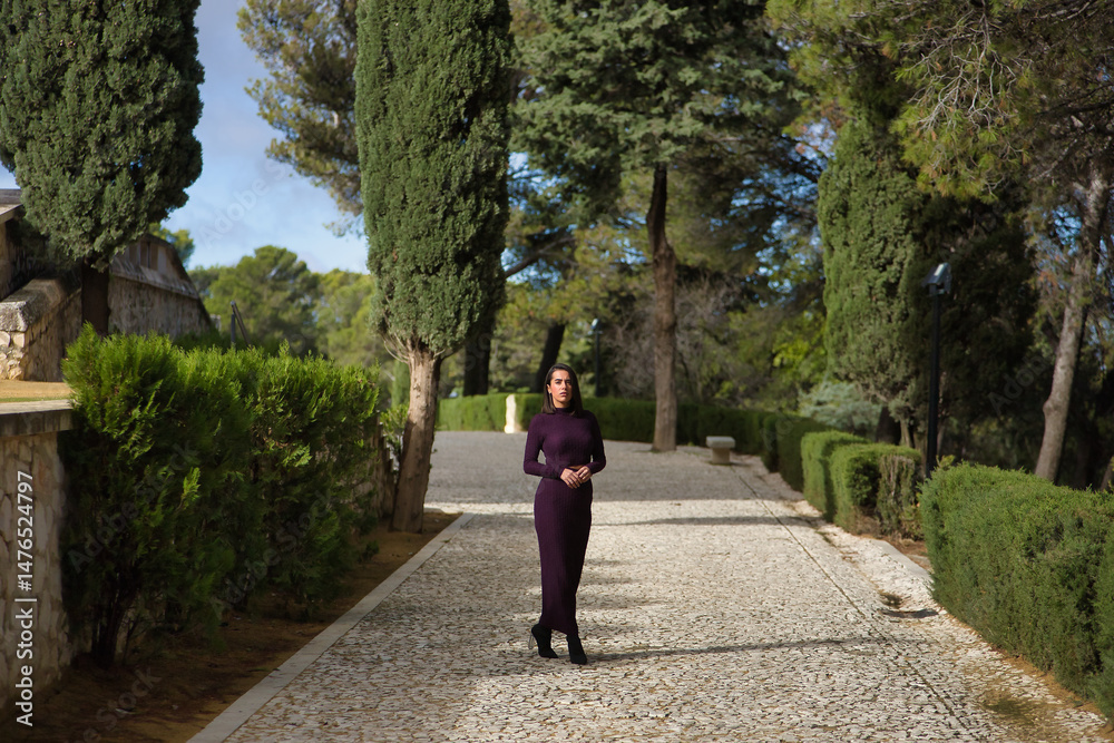Naklejka premium Young, beautiful, brunette, Hispanic woman, wearing a purple dress and high heels, walking alone on a stone path surrounded by cypress trees. Concept beauty, fashion, trendy.