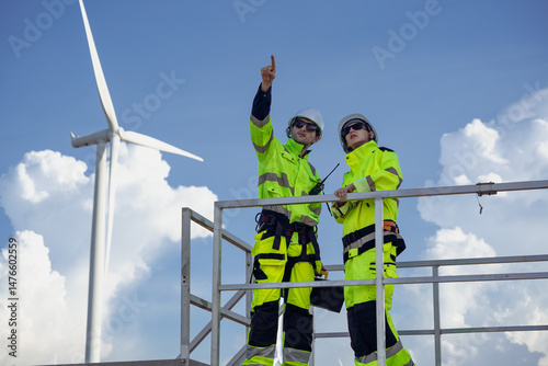 Maintenance engineer team standing at windmills at wind turbine farm. Group of people wear safety helmet and uniform working at alternative renewable energy wind station. Sustainable energy technology