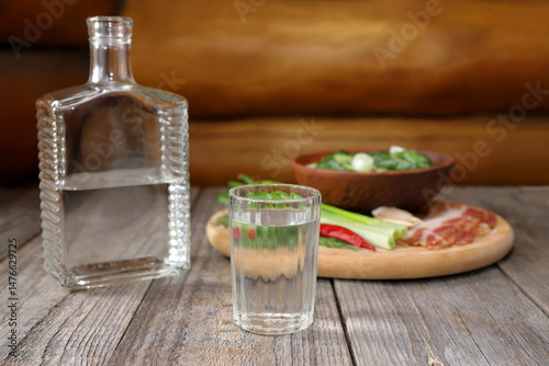 On a wooden table against the log wall there is a shot of vodka in front of a glass pitcher next to a wooden tray with a snack of vegetables, bacon and a clay bowl of pickled cucumbers. 