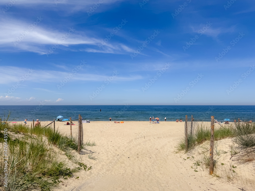 A sunny beach scene with a sandy path leading to the Baltic Sea under a partly cloudy blue sky, with scattered people enjoying the shoreline. A sunny beach scene with a sandy path leading to the Baltic Sea under a partly cloudy blue sky, with scattered people enjoying the shoreline.