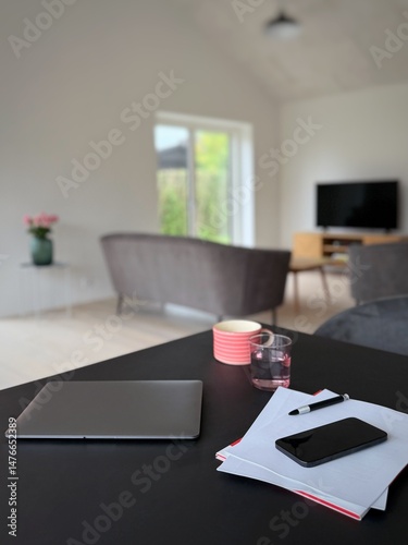 Close-up of a closed laptop computer, paperwork, pen, mobile phone and drinks on a dining room table