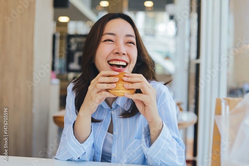A cheerful young Asian woman is smiling brightly while holding a burger with both hands, ready to take a bite. She is enjoying her meal at a modern cafe or restaurant.