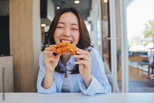 A joyful young Asian woman enjoying a piece of fried chicken at a modern restaurant. She laughs wholeheartedly, holding the crispy food.