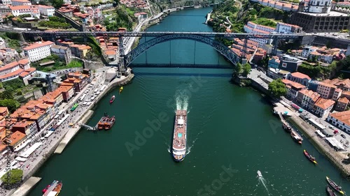 Aerial of a river cruise ship sailing beneath the iconic Dom Luís I Bridge in Porto, surrounded by historic buildings and bustling riverbanks