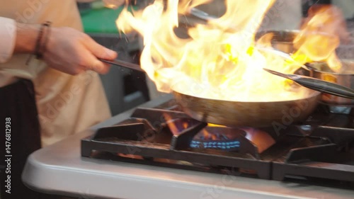 Professional chef working in a commercial kitchen preparing a meal with steam rising from the cooking pans and other kitchen equipment visible The chef is focused on the cooking process, pouring sauce