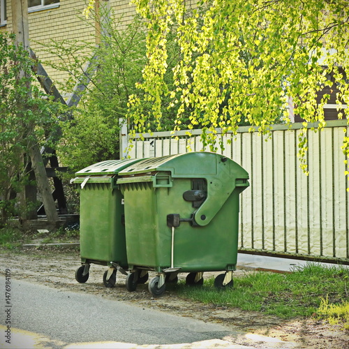 green trash containers in sunlit street near fence with birch tree branches and greenery