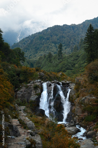 A serene glacier melted water's fall cascades down rocky cliffs surrounded by dense green forest and misty mountains during autumn season